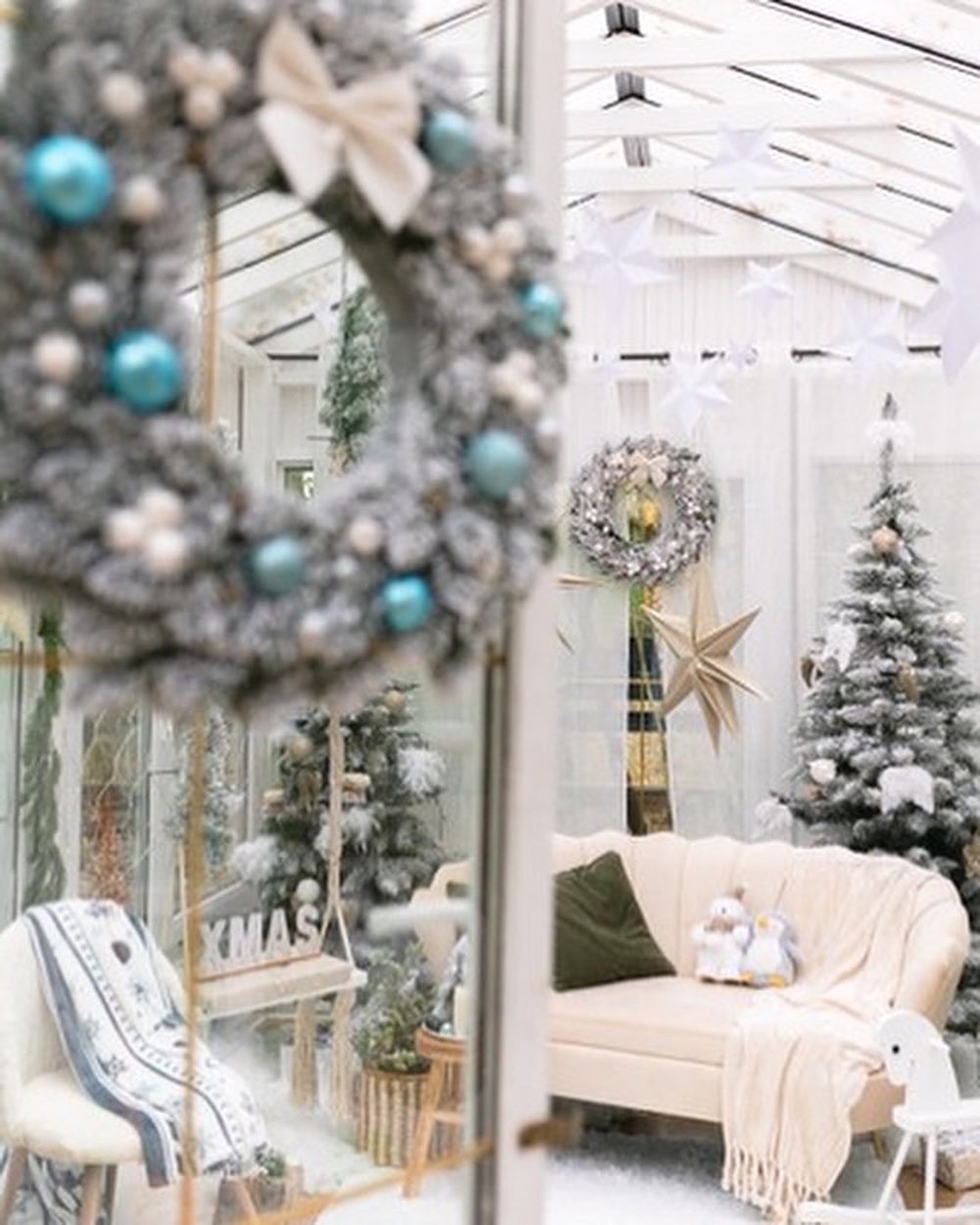 Dekoracja wnętrza w stylu zimowym: ośnieżony wieniec z błękitnymi bombkami w kadrze, choinki, sofa z poduszką i pluszowymi pingwinami, oraz wiszące gwiazdy z papieru.