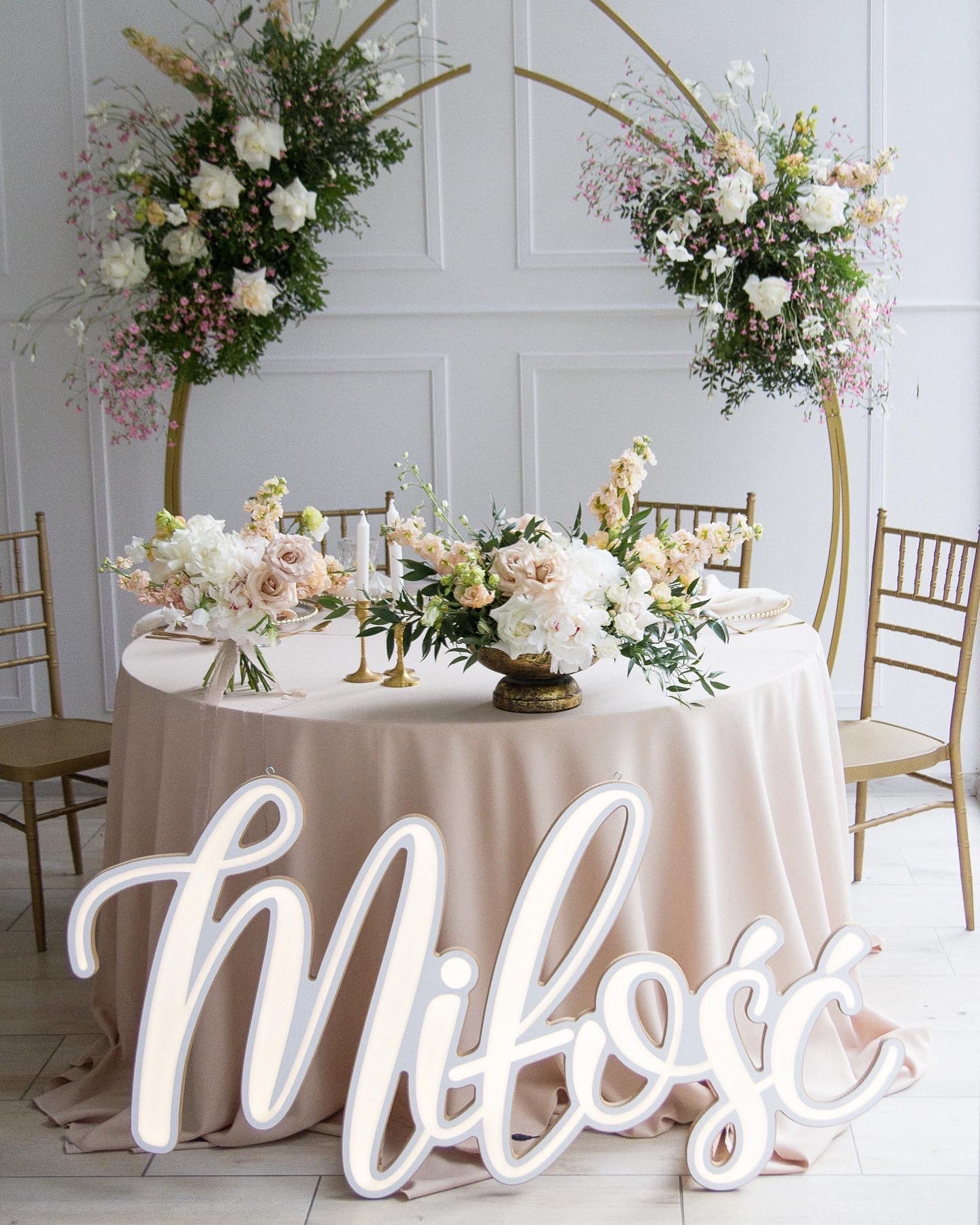 Dekoracja stołu weselnego z kwiatowymi kompozycjami w odcieniach bieli i różu, złotymi świecznikami i napisem 'Miłość' w dekoracyjnej czcionce.