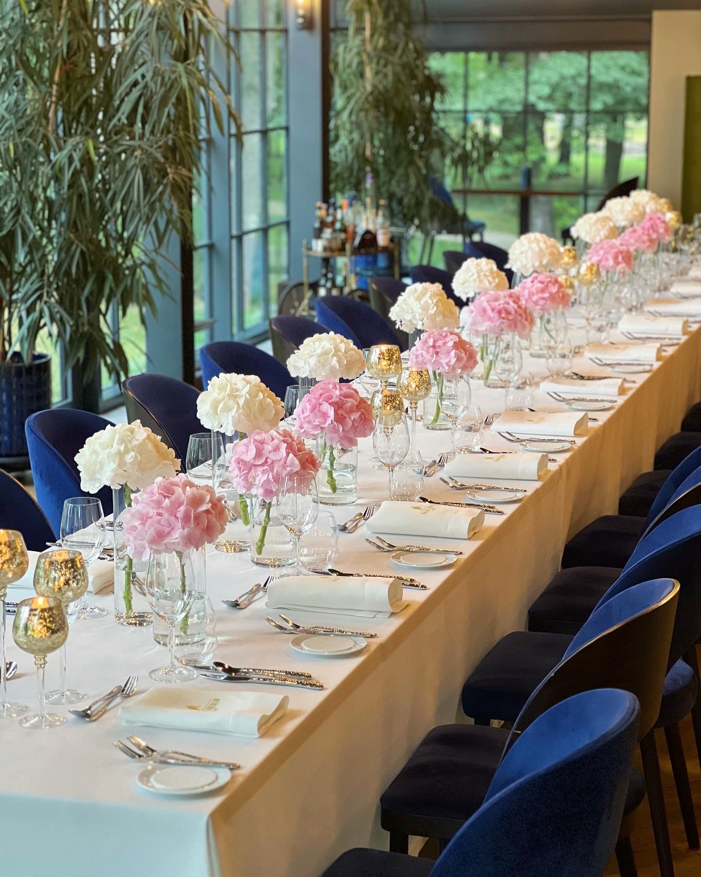 Długi stół udekorowany na przyjęcie, z białym obrusem, kwiatami hortensji w odcieniach różu i bieli, elegancką zastawą i złotymi świecznikami, otoczony granatowymi krzesłami, z widokiem na zieleń...