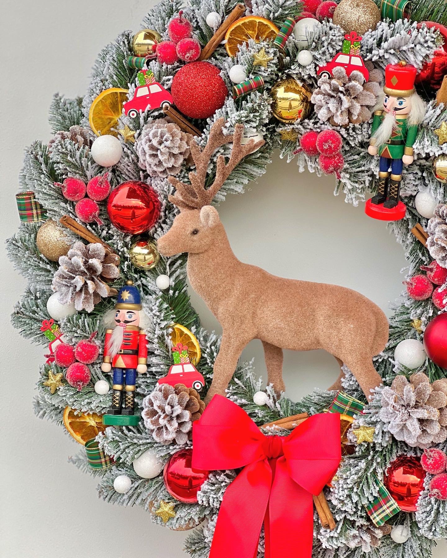 Świąteczny wieniec z pluszowym reniferem w centrum, ozdobiony bombkami, szyszkami, suszonymi pomarańczami, czerwonymi koralikami, drewnianymi dziadkami do orzechów i czerwoną kokardą na dole...