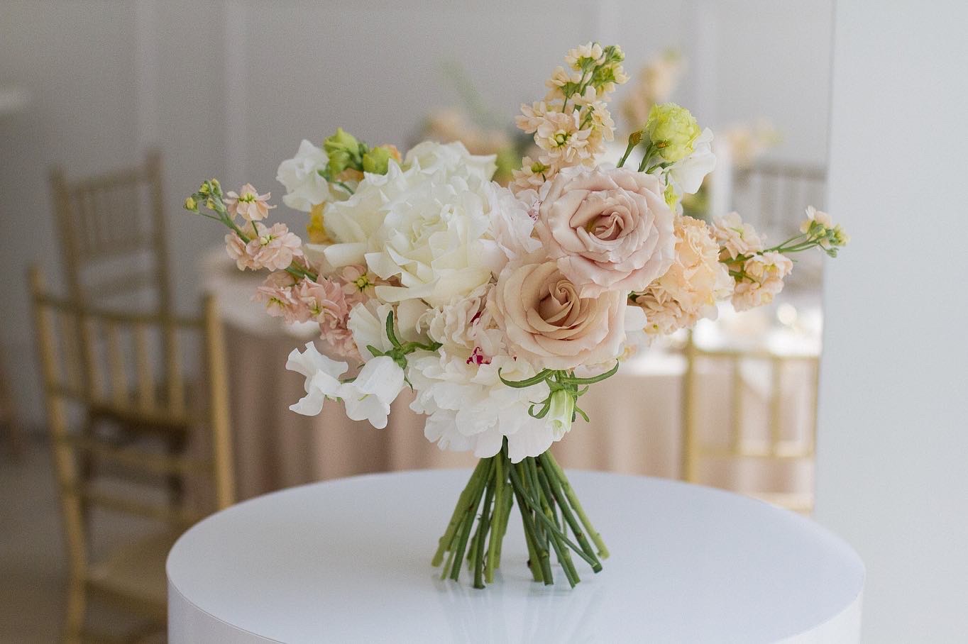 Bukiet ślubny z róż, piwonii i groszku pachnącego w odcieniach bieli, różu i kremu na białym okrągłym stoliku, w tle złote krzesła i stół przykryty beżowym obrusem.