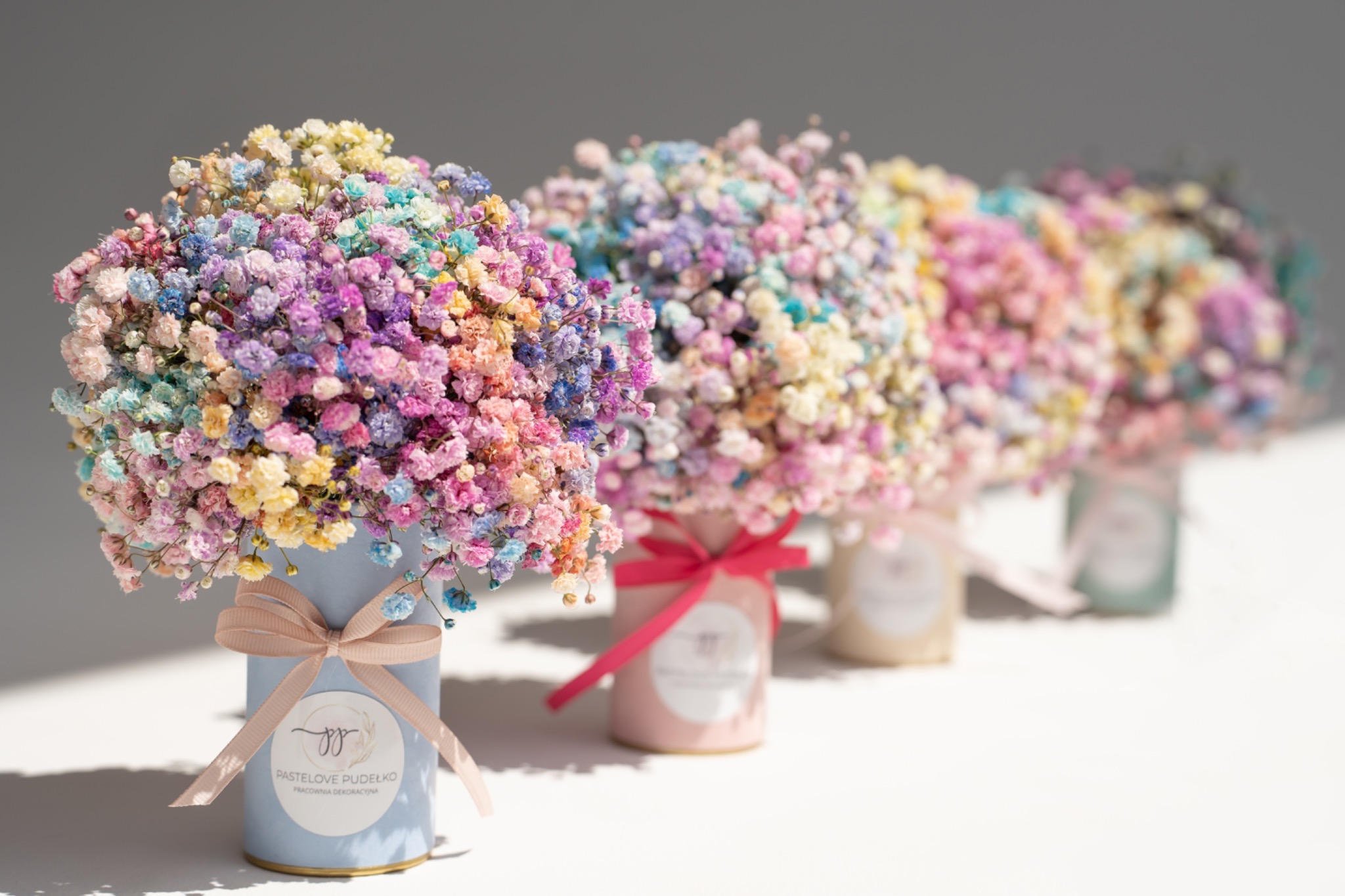 Rząd pastelowych pudełek z wielobarwnymi bukietami gipsówki, każde pudełko przewiązane wstążką w dopasowanym kolorze, na jasnym tle.