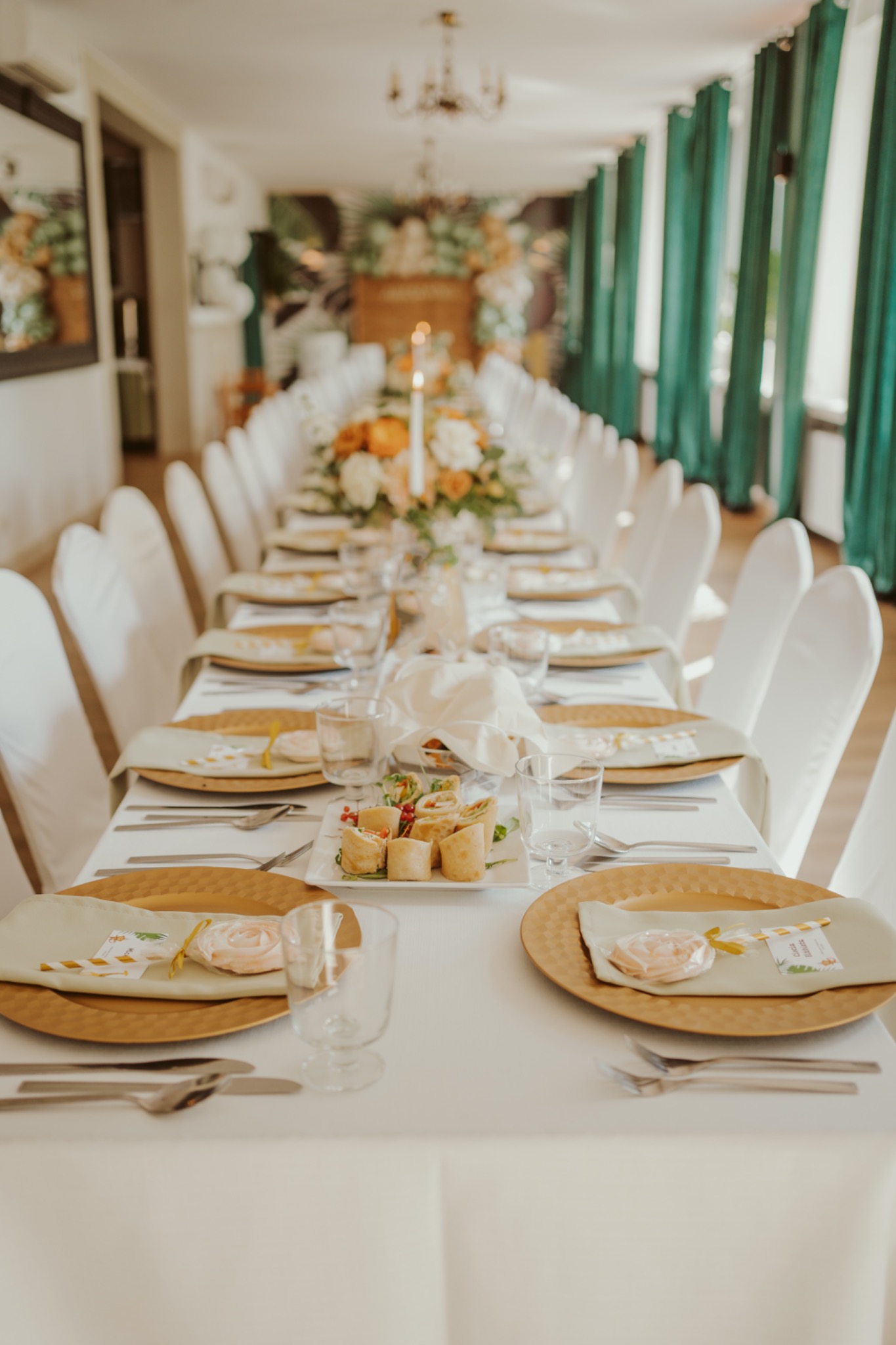 Długi stół z białym obrusem, nakryty złotymi talerzami, jasnymi serwetkami, sztućcami i szklankami, udekorowany kwiatami i świecami, z zielonymi zasłonami w tle.