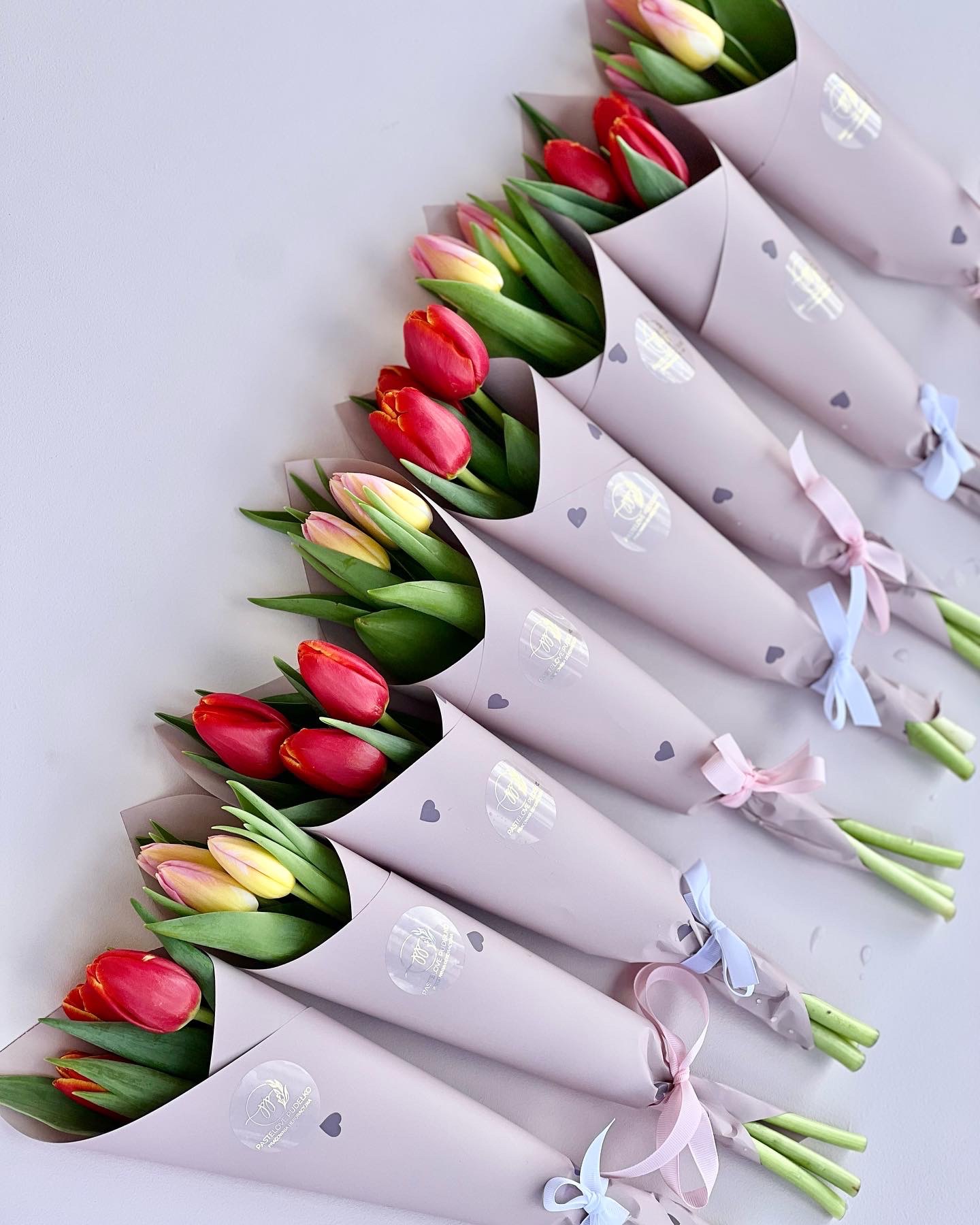 Rząd siedmiu bukietów tulipanów w pastelowym papierze z kokardkami, widok z góry, kompozycja kwiatowa.