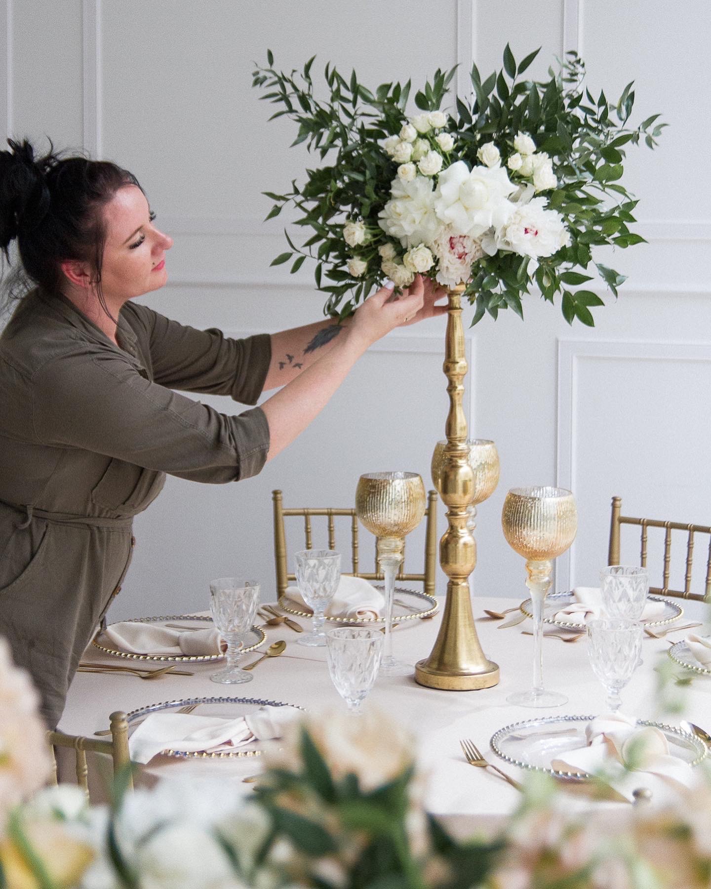 Kobieta w kombinezonie dogląda kompozycji z białych kwiatów w wysokim, złotym wazonie na stole z elegancką zastawą