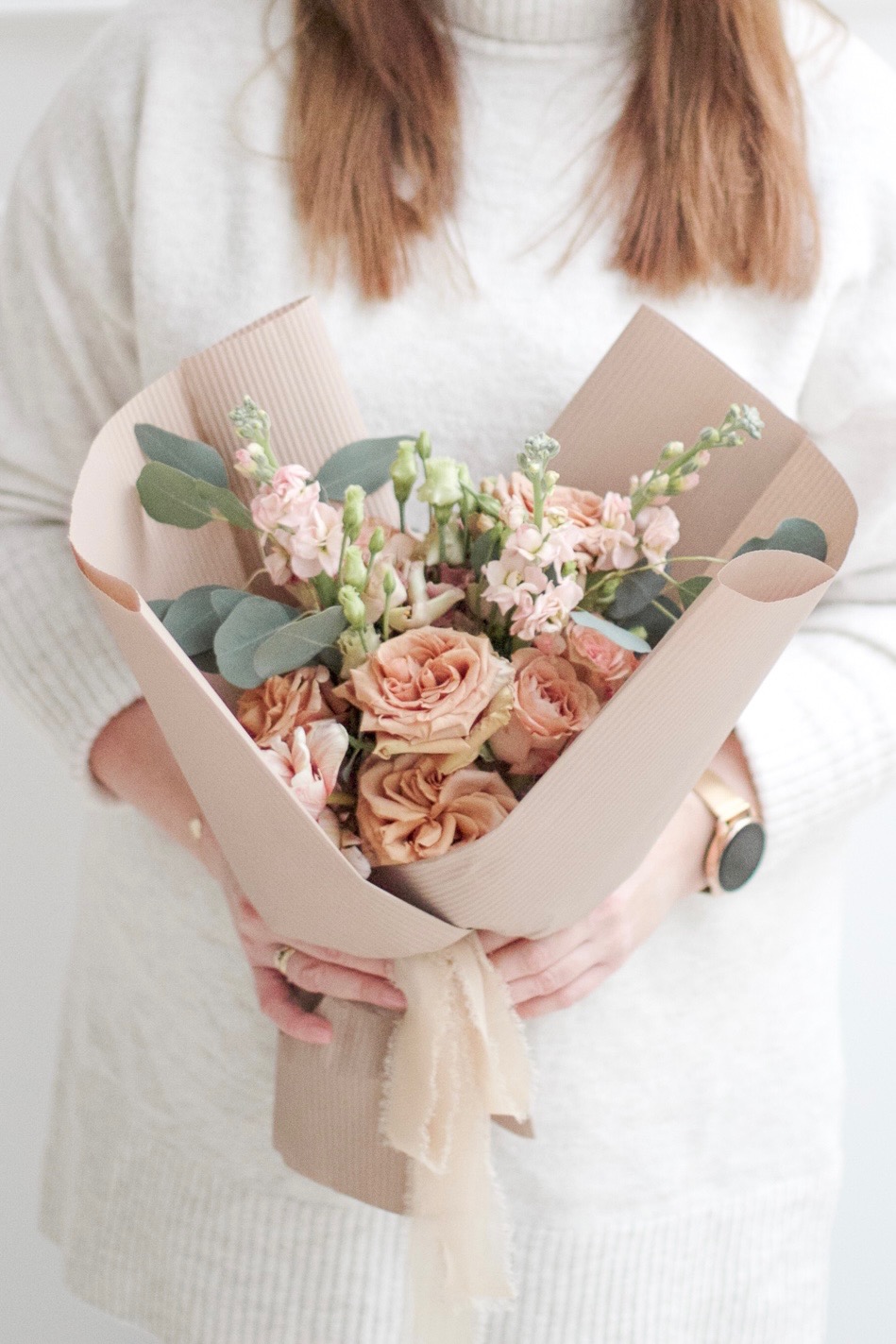 Bukiet brzoskwiniowych róż i białych kwiatów w papierowym opakowaniu z beżową wstążką, trzymany przez osobę w białym swetrze i złotym zegarku.
