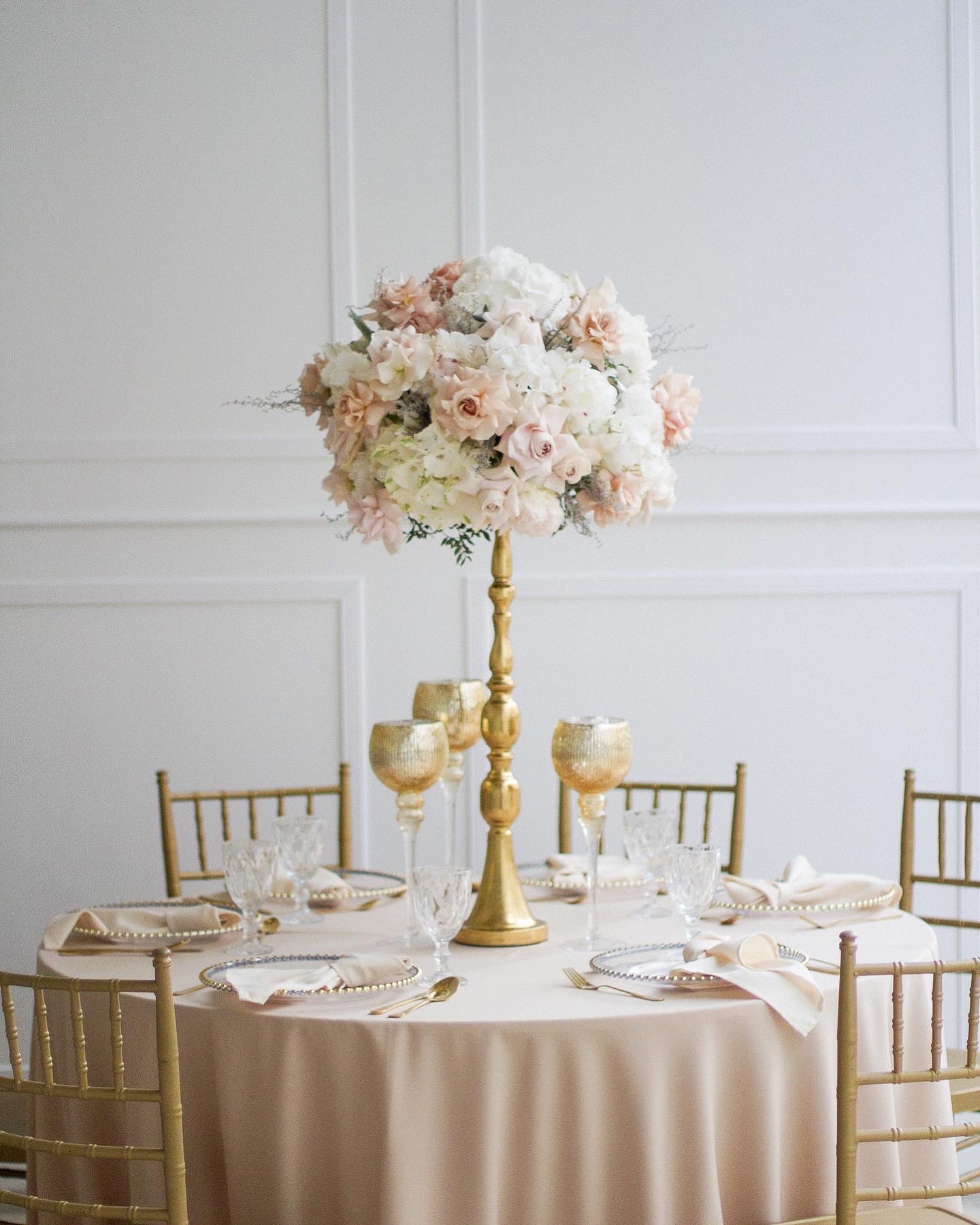 Uroczysty stół z okrągłym obrusem w kolorze brzoskwini, złote krzesła, wysoki złoty świecznik z kompozycją kwiatową w odcieniach bieli i różu, elegancka zastawa stołowa ze złotymi akcentami...