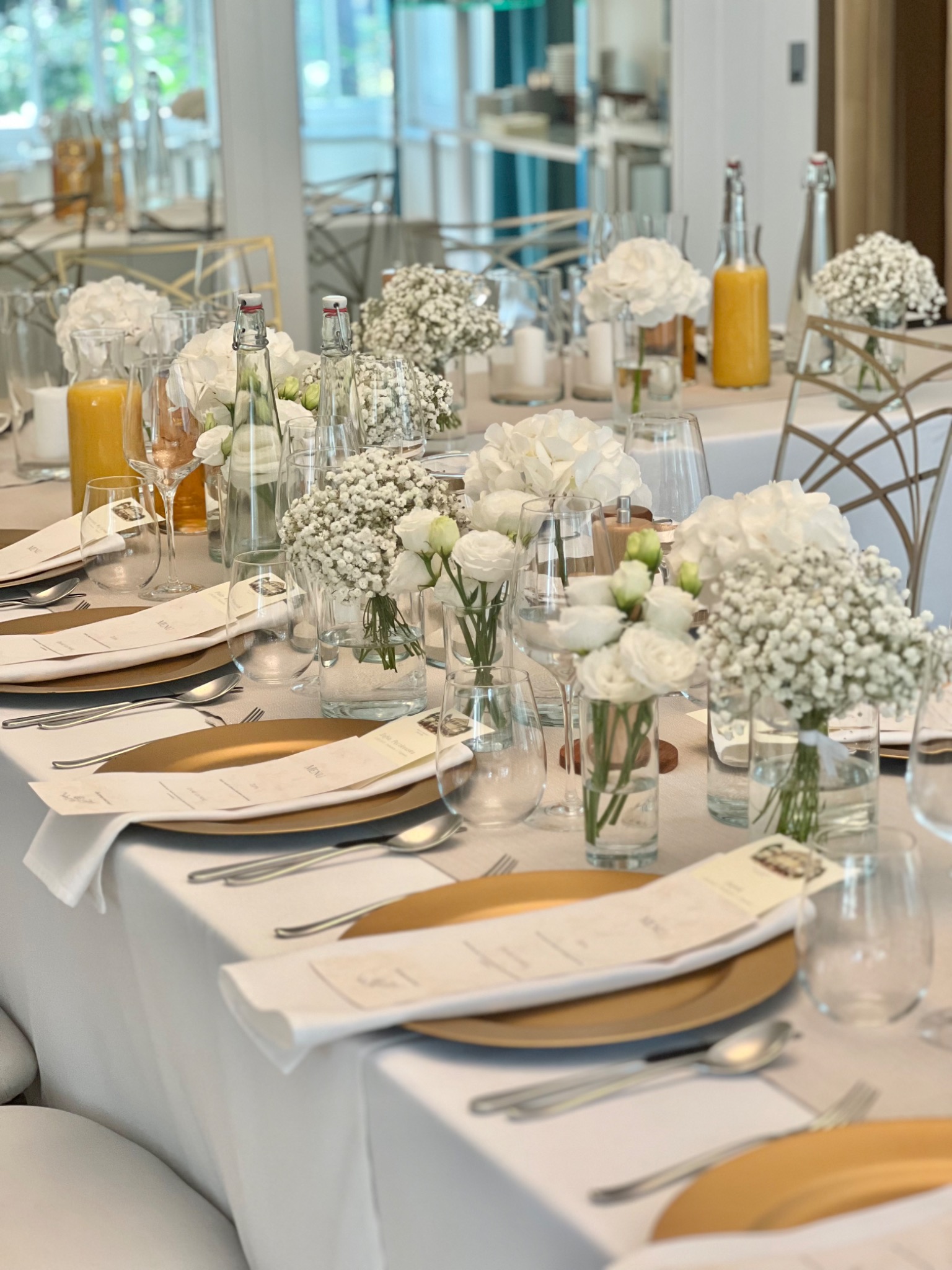 Elegancko nakryty stół z białym obrusem, złotymi talerzami, menu, szklankami, butelkami z wodą i sokiem oraz kompozycjami z białych kwiatów, przygotowany na uroczystość.