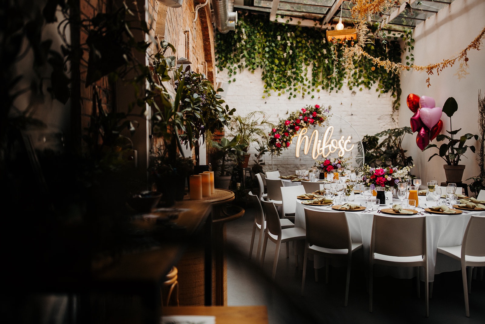 Uroczysta dekoracja sali z okrągłymi stołami, białymi krzesłami, kwiatowymi kompozycjami, balonami w kształcie serc i neonowym napisem 'Miłość' na tle ściany z cegły i zieleni.