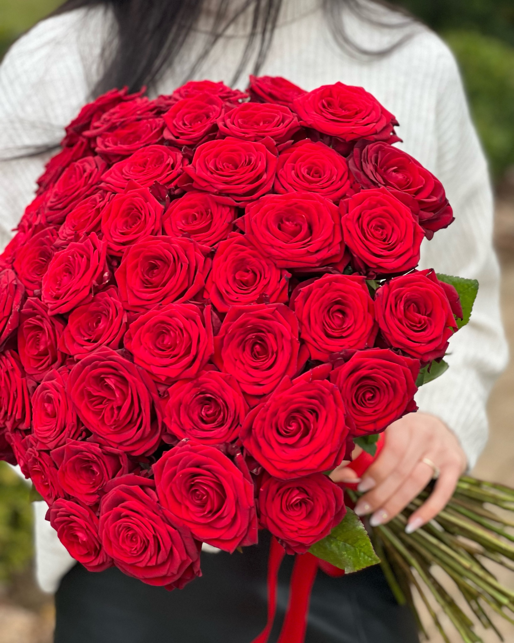 Kobieta w białym swetrze trzyma bukiet intensywnie czerwonych róż, związanych czerwoną wstążką, z widocznym pierścionkiem na palcu.