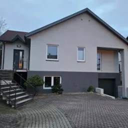 Dwukondygnacyjny dom z szarą elewacją, widoczne schody wejściowe z metalową barierką, kostka brukowa na podjeździe i sąsiedni budynek w stanie surowym.