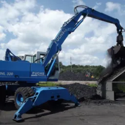 Niebieska koparka Fuchs MHL 320 z chwytakiem ładuje opał na rampę. W tle hałdy węgla i błękitne niebo z chmurami.