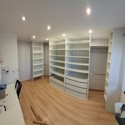 HR-Home Łukasz Ślusarek - Przestronna garderoba z białymi, modułowymi szafami z półkami i szufladami, jasne drewniane panele podłogowe, widok z perspektywy szerokokątnej, oświetlenie punktowe sufitowe.