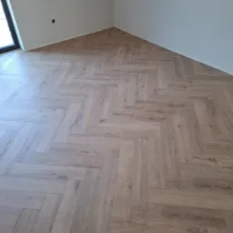 Jasne panele ułożone w jodełkę francuską w rogu pokoju, z widocznym gniazdkiem elektrycznym na ścianie i oknem po lewej stronie.