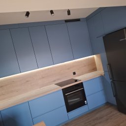 HR-Home Łukasz Ślusarek - Niebieska kuchnia z drewnianym blatem i oświetleniem LED. Nowoczesny design, czarny piekarnik i lodówka. Ukośne ujęcie podkreśla głębię i przestrzeń wnętrza.