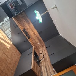 HR-Home Łukasz Ślusarek - Nowoczesna kuchnia na wymiar z szarymi szafkami i drewnianym blatem, oświetlona lampą z zielonym abażurem. Widoczny fragment płyty grzewczej i zlewozmywak.