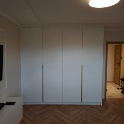 HR-Home Łukasz Ślusarek - Biała, czterodrzwiowa szafa wnękowa z długimi, smukłymi uchwytami, na tle parkietu ułożonego w jodełkę. Widoczne drzwi do innego pomieszczenia.