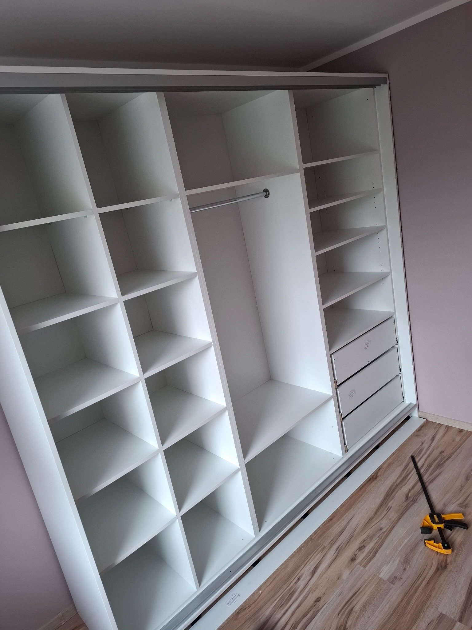 Wnętrze białej, przesuwnej szafy wnękowej z półkami, drążkiem na ubrania i trzema szufladami, widoczna klamra stolarska na podłodze z paneli.