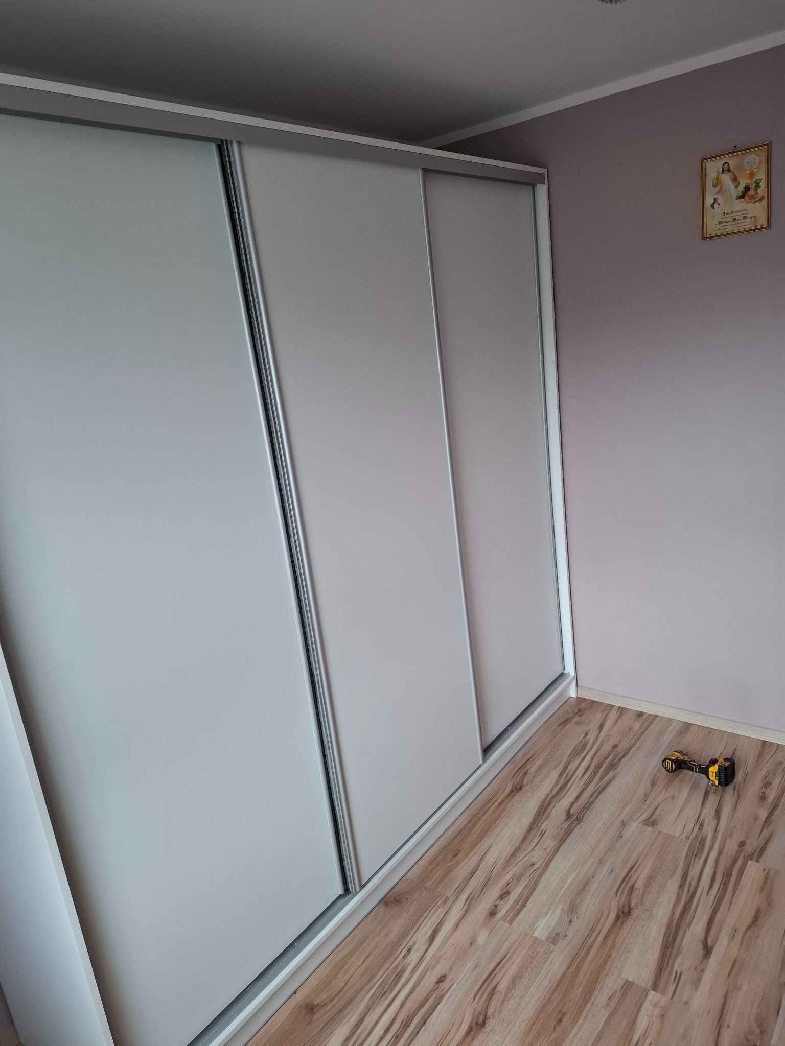 Jasna szafa przesuwna z trzema drzwiami na drewnianej podłodze, w rogu pomieszczenia w trakcie montażu, obok leży wkrętarka akumulatorowa.