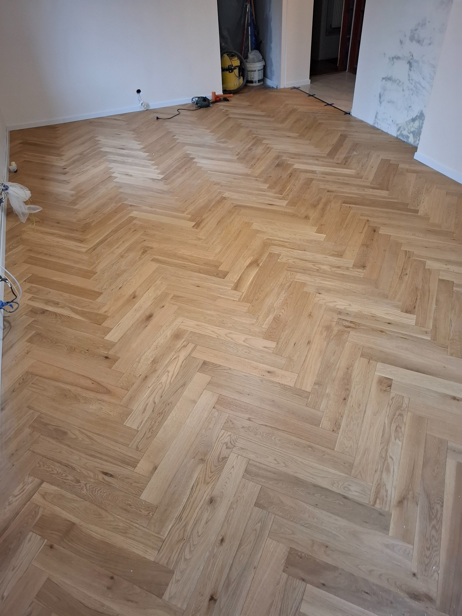 Jasny parkiet ułożony we wzór jodełki francuskiej, widoczne narzędzia i materiały budowlane w tle, pokój po remoncie w trakcie wykańczania.