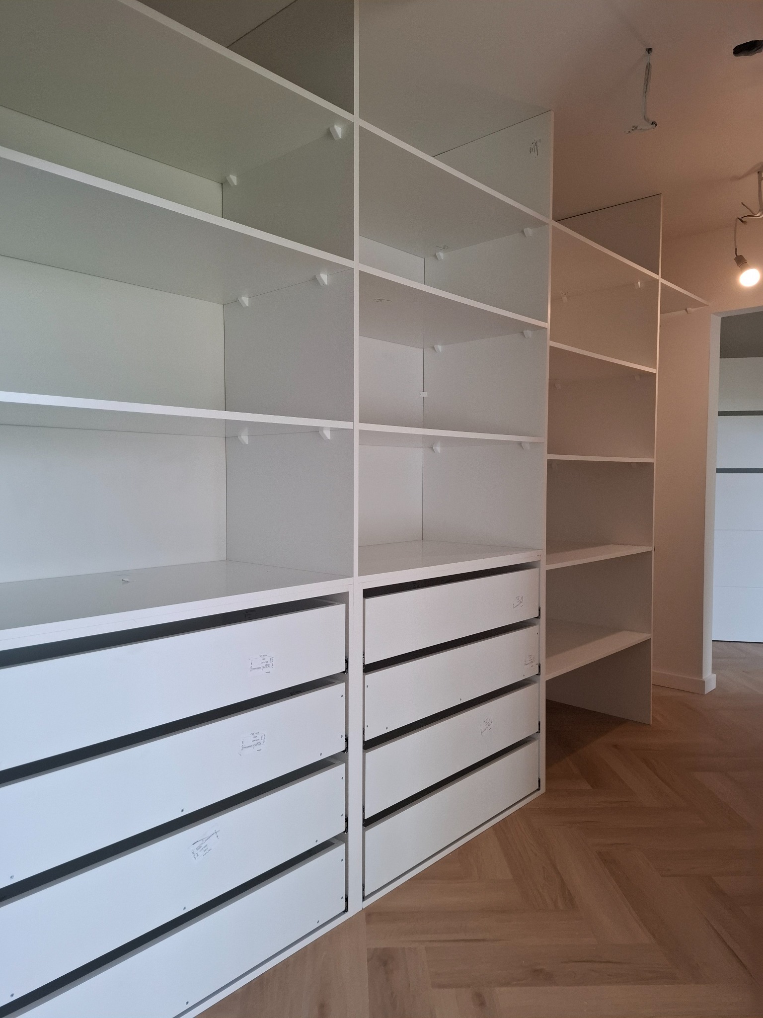 Biała, nowoczesna garderoba z szufladami i półkami, zamontowana w pomieszczeniu z parkietem ułożonym w jodełkę, widoczne punkty mocowania na ścianie.