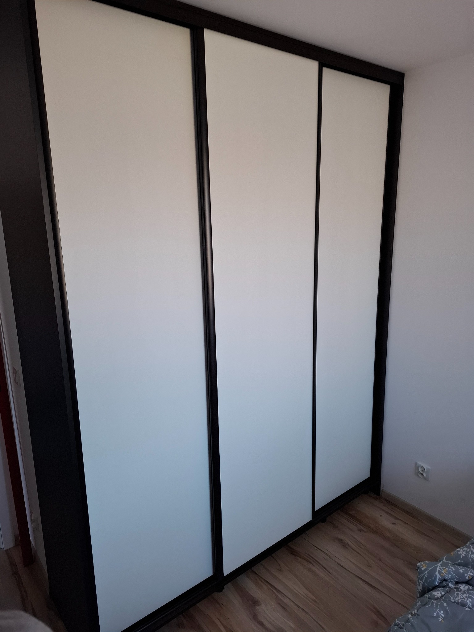 Nowoczesna szafa przesuwna z trzema białymi frontami i ciemnymi obramowaniami, widoczna z boku w sypialni na tle białej ściany i drewnianej podłogi.