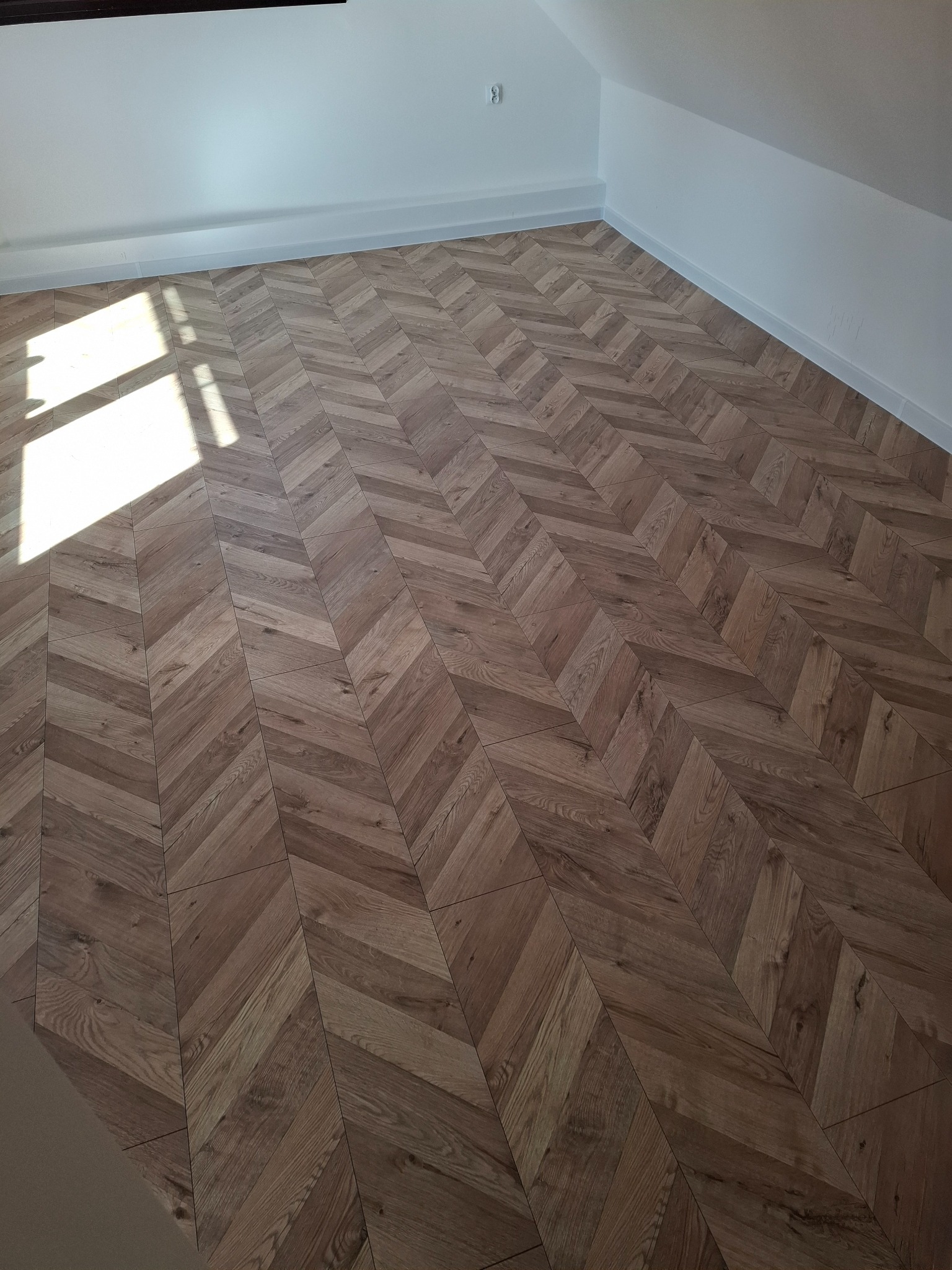 Jasnobrązowa podłoga ułożona w jodełkę w nowym pokoju, widoczne naturalne światło z okna, białe listwy przypodłogowe.