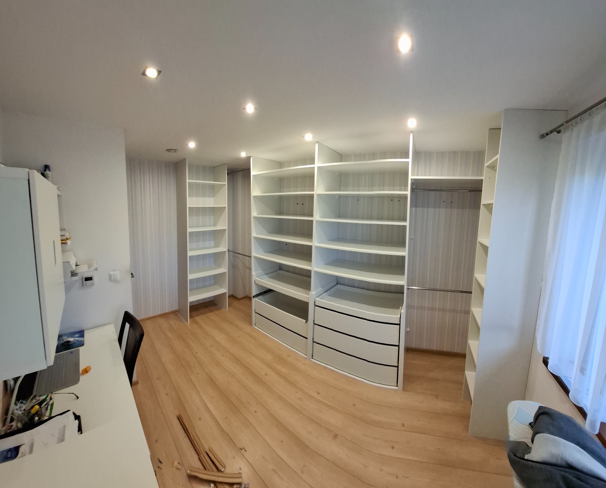 Przestronna garderoba z białymi, modułowymi szafami z półkami i szufladami, jasne drewniane panele podłogowe, widok z perspektywy szerokokątnej, oświetlenie punktowe sufitowe.