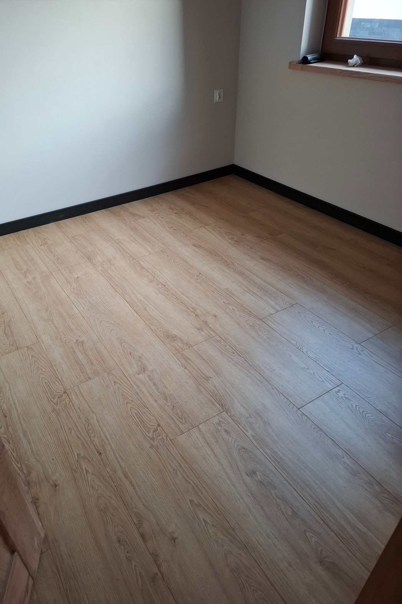 Jasne panele podłogowe z widoczną strukturą drewna, ułożone w pomieszczeniu z białymi ścianami i czarnymi listwami przypodłogowymi, oświetlone naturalnym światłem wpadającym przez okno.