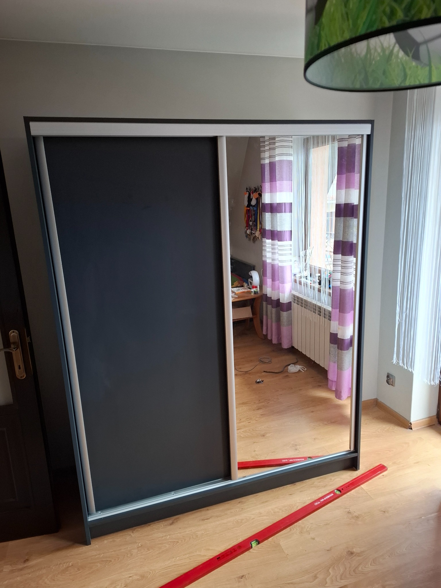Ciemnoszara szafa wnękowa z przesuwnymi drzwiami z lustrem, poziomica na podłodze, widoczny fragment pokoju z grzejnikiem i zasłonami.