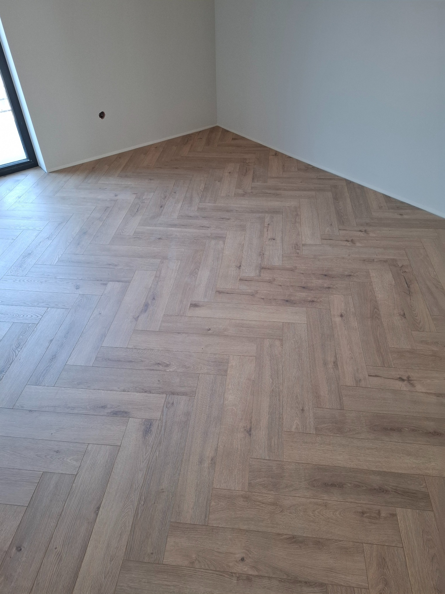 Jasne panele ułożone w jodełkę francuską w rogu pokoju, z widocznym gniazdkiem elektrycznym na ścianie i oknem po lewej stronie.