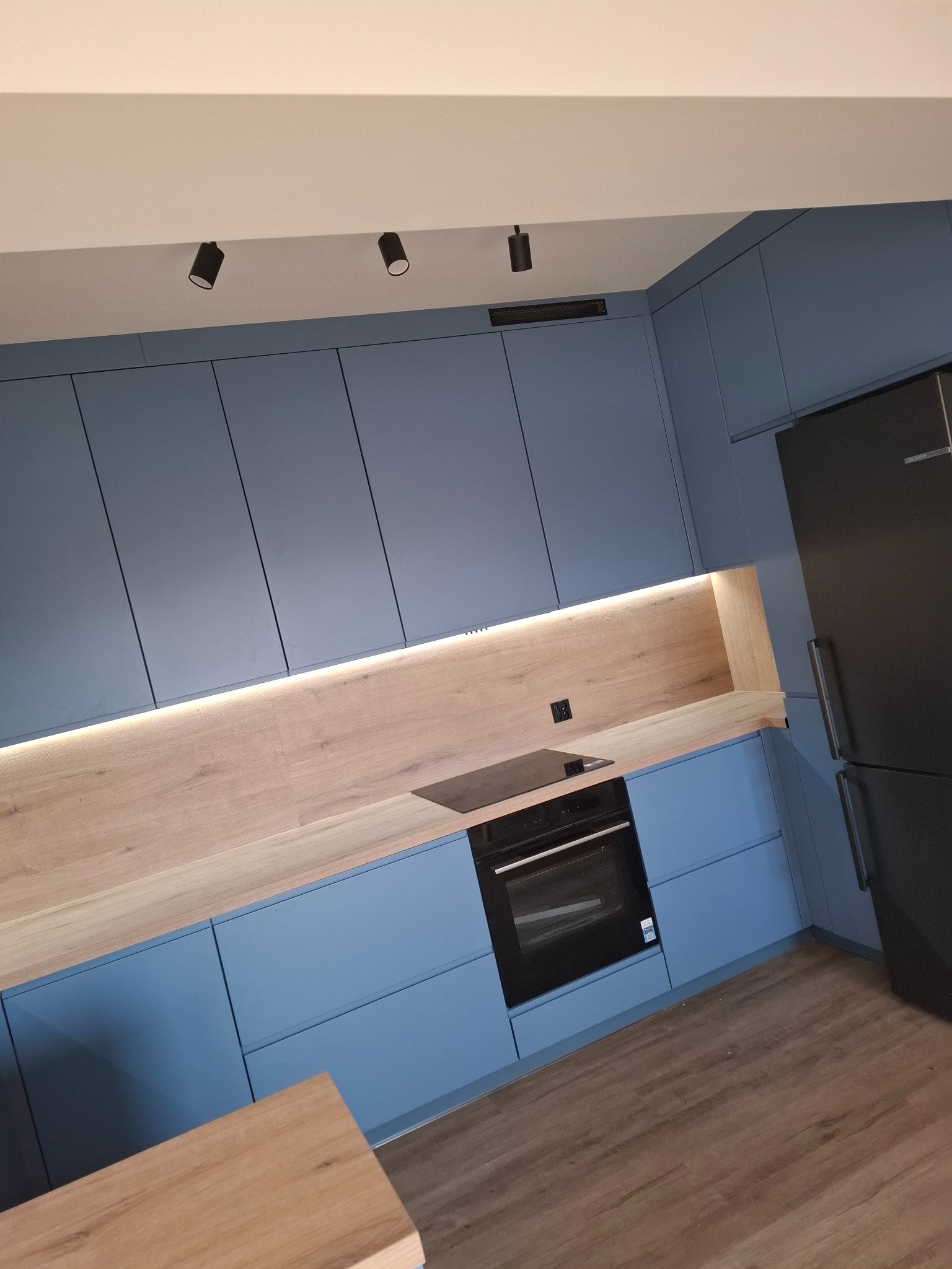 Niebieska kuchnia z drewnianym blatem i oświetleniem LED. Nowoczesny design, czarny piekarnik i lodówka. Ukośne ujęcie podkreśla głębię i przestrzeń wnętrza.