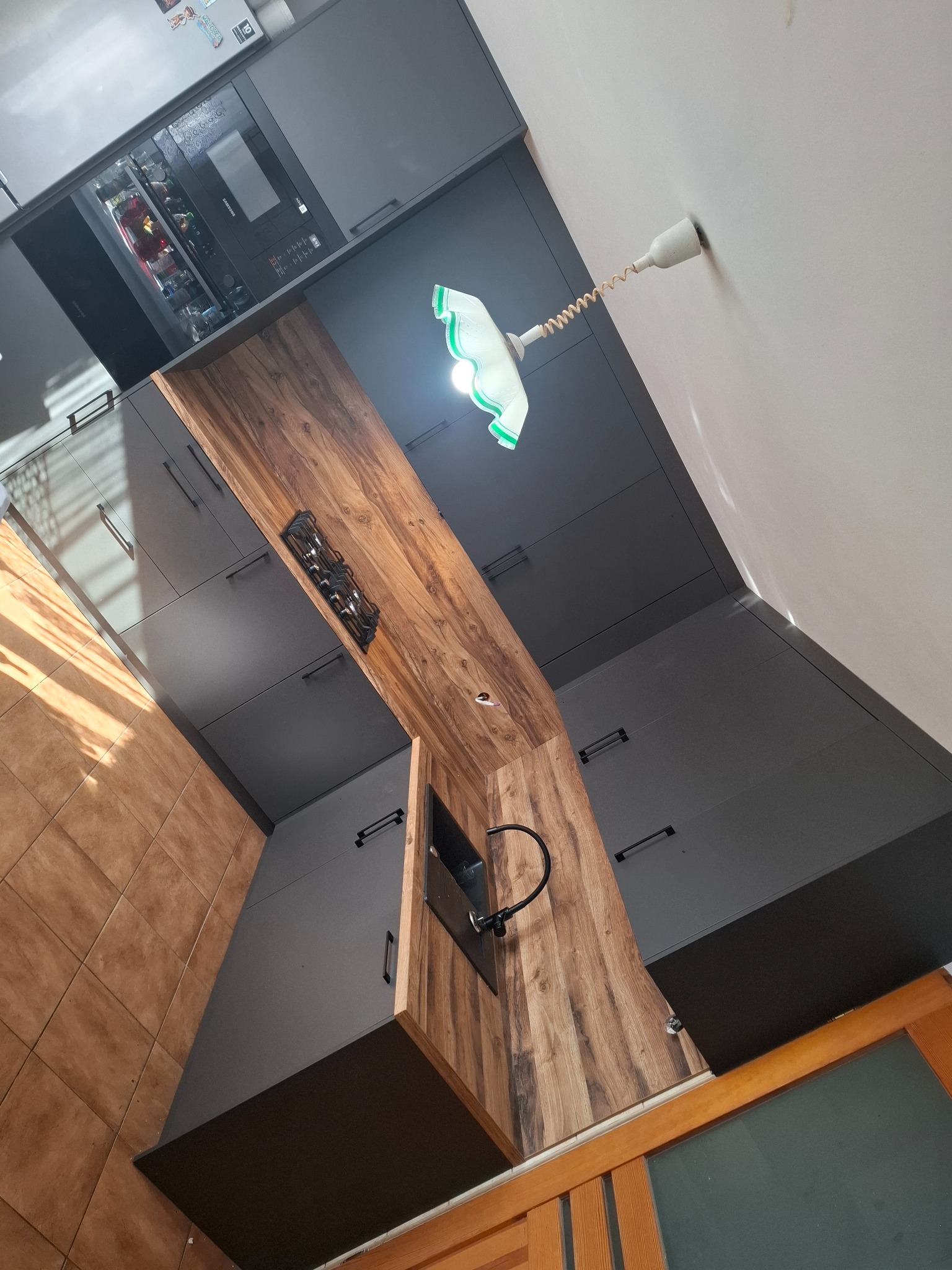 Nowoczesna kuchnia na wymiar z szarymi szafkami i drewnianym blatem, oświetlona lampą z zielonym abażurem. Widoczny fragment płyty grzewczej i zlewozmywak.