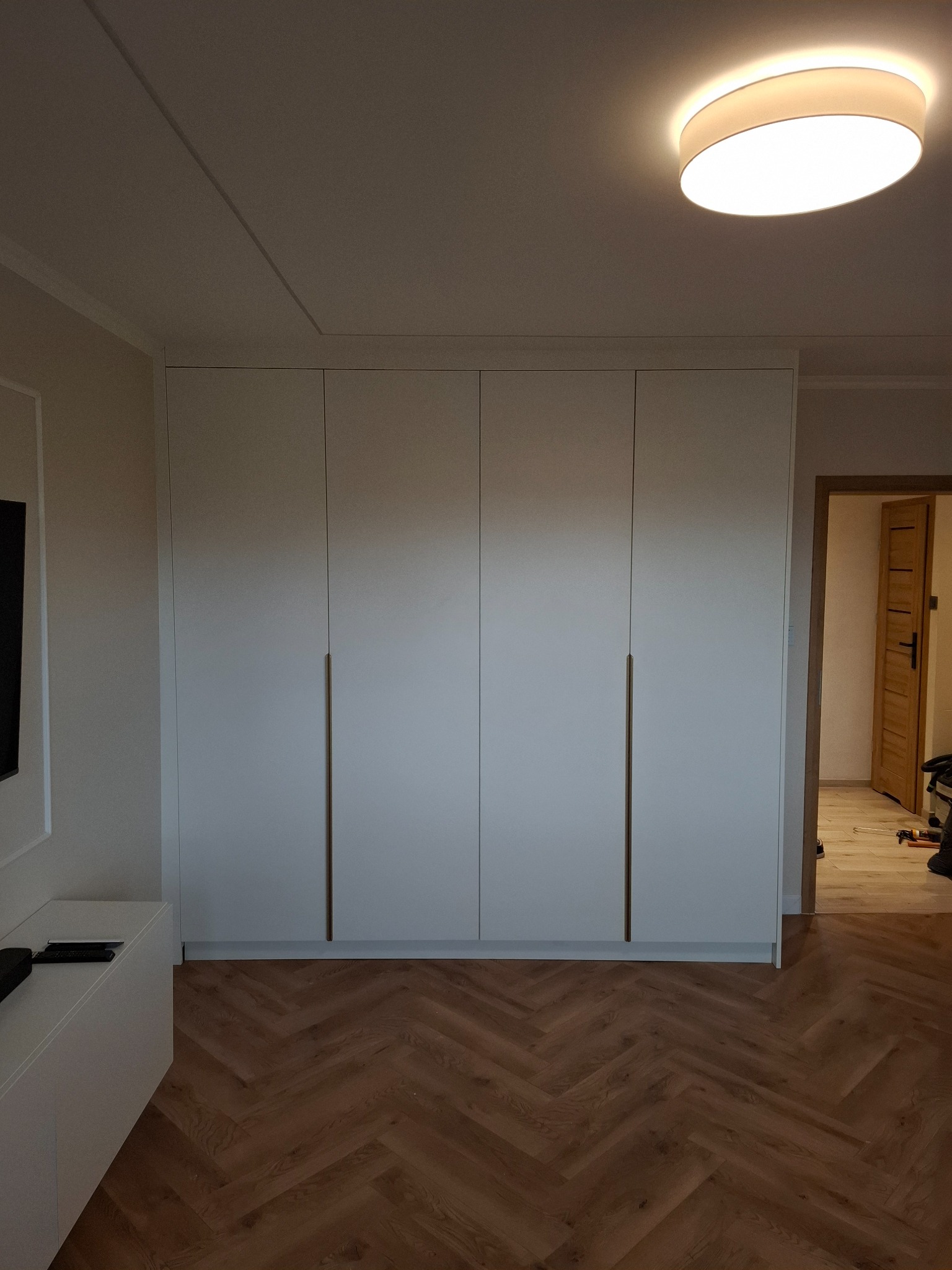 Biała, czterodrzwiowa szafa wnękowa z długimi, smukłymi uchwytami, na tle parkietu ułożonego w jodełkę. Widoczne drzwi do innego pomieszczenia.