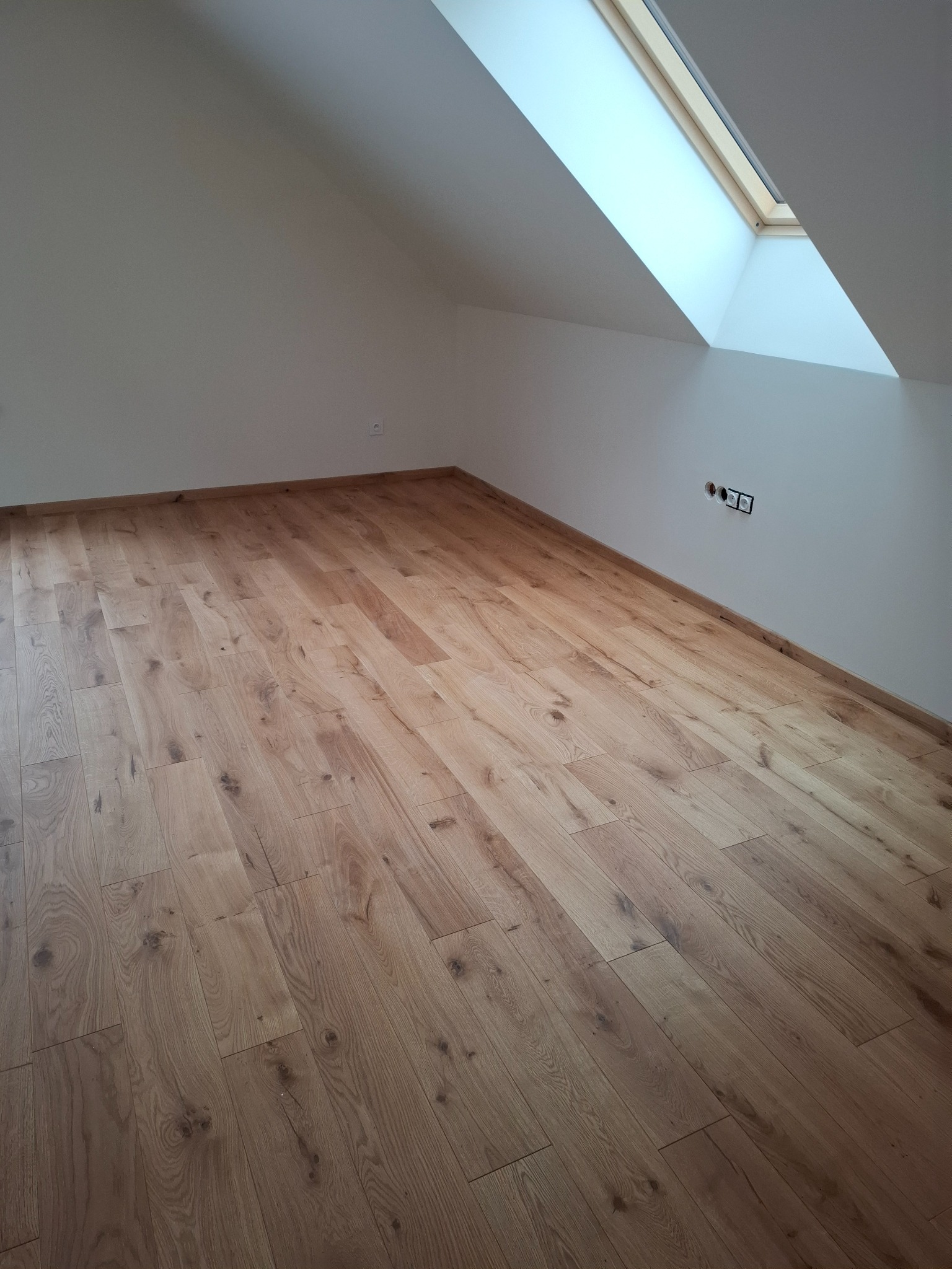 Jasne panele podłogowe w pokoju na poddaszu z oknem dachowym. Widoczne gniazdka elektryczne na białej ścianie. Ukośne ściany i skosy sufitu.