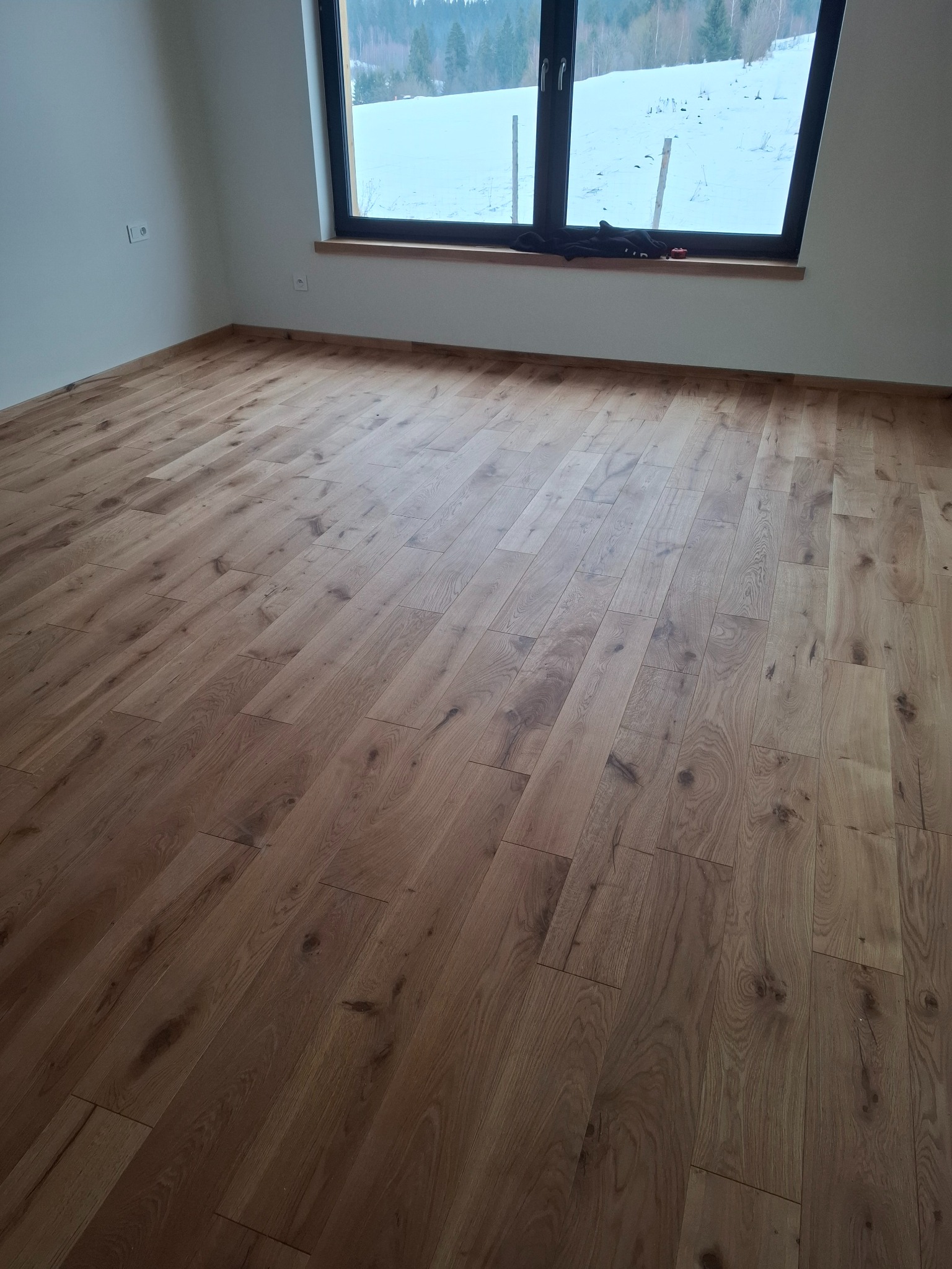 Ukończona podłoga z paneli drewnianych w jasnym odcieniu, widok z perspektywy, okno z widokiem na zaśnieżony krajobraz górski, minimalistyczne wnętrze.