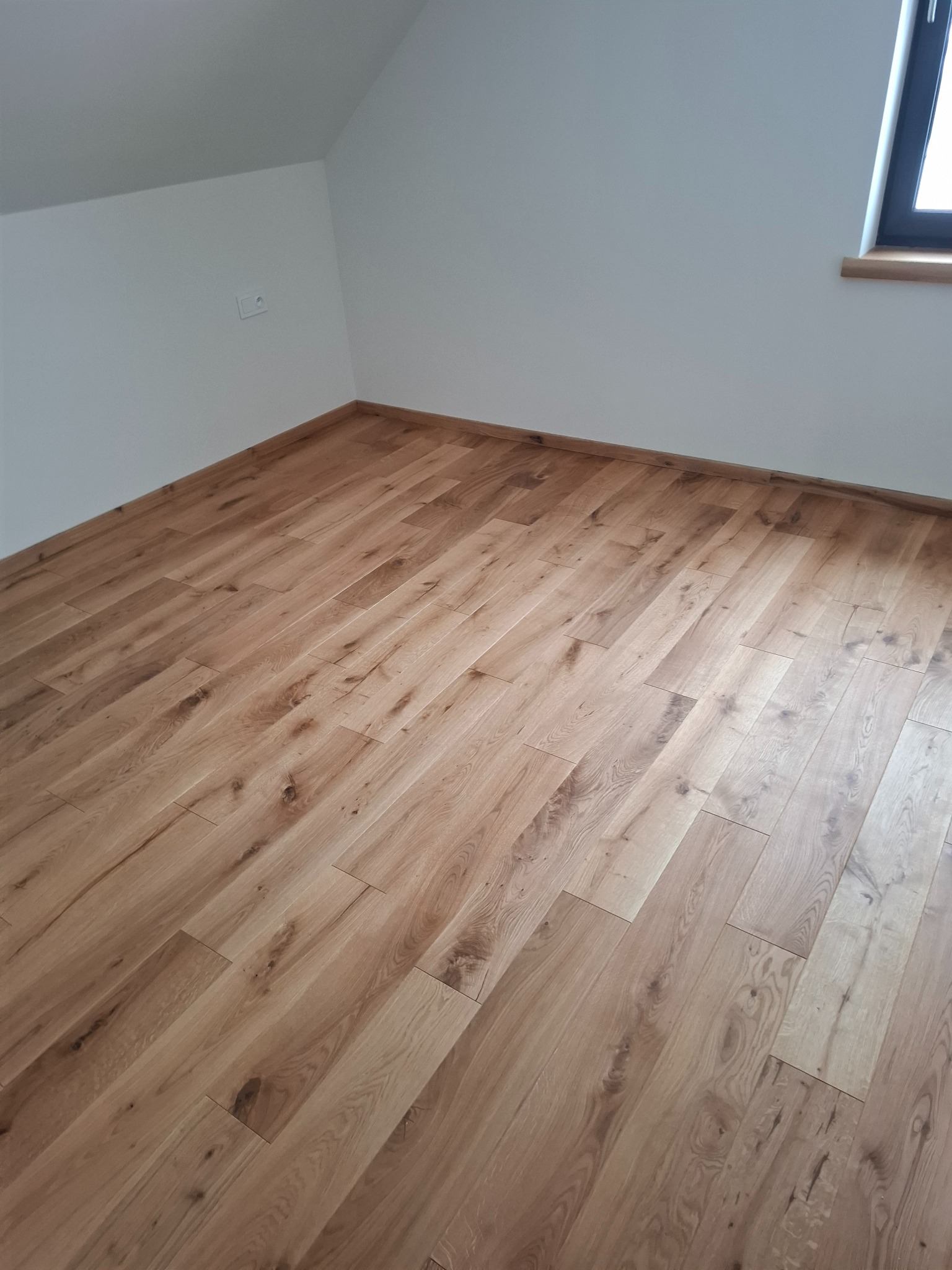 Jasne panele podłogowe ułożone w pokoju na poddaszu, widoczne okno i gniazdko elektryczne. Naturalne oświetlenie podkreśla strukturę drewna.