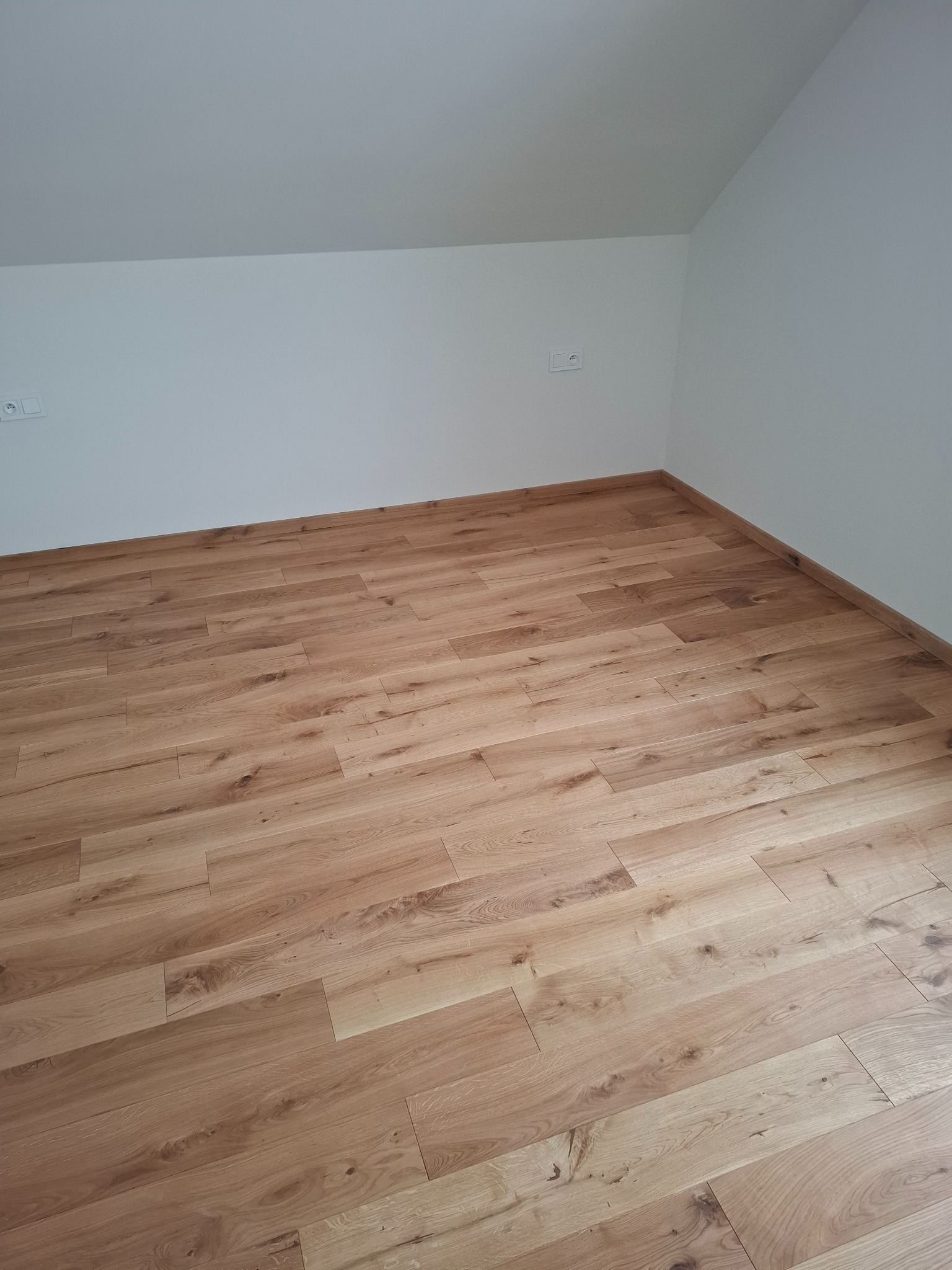 Jasne panele podłogowe z wyraźnym rysunkiem drewna, ułożone w nowym pokoju z białymi ścianami i skośnym sufitem. Widoczne gniazdka elektryczne.