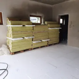 Stos płyt izolacyjnych z wełny mineralnej na paletach w surowym wnętrzu, widoczne okno i przygotowana podłoga z płyt styropianowych.