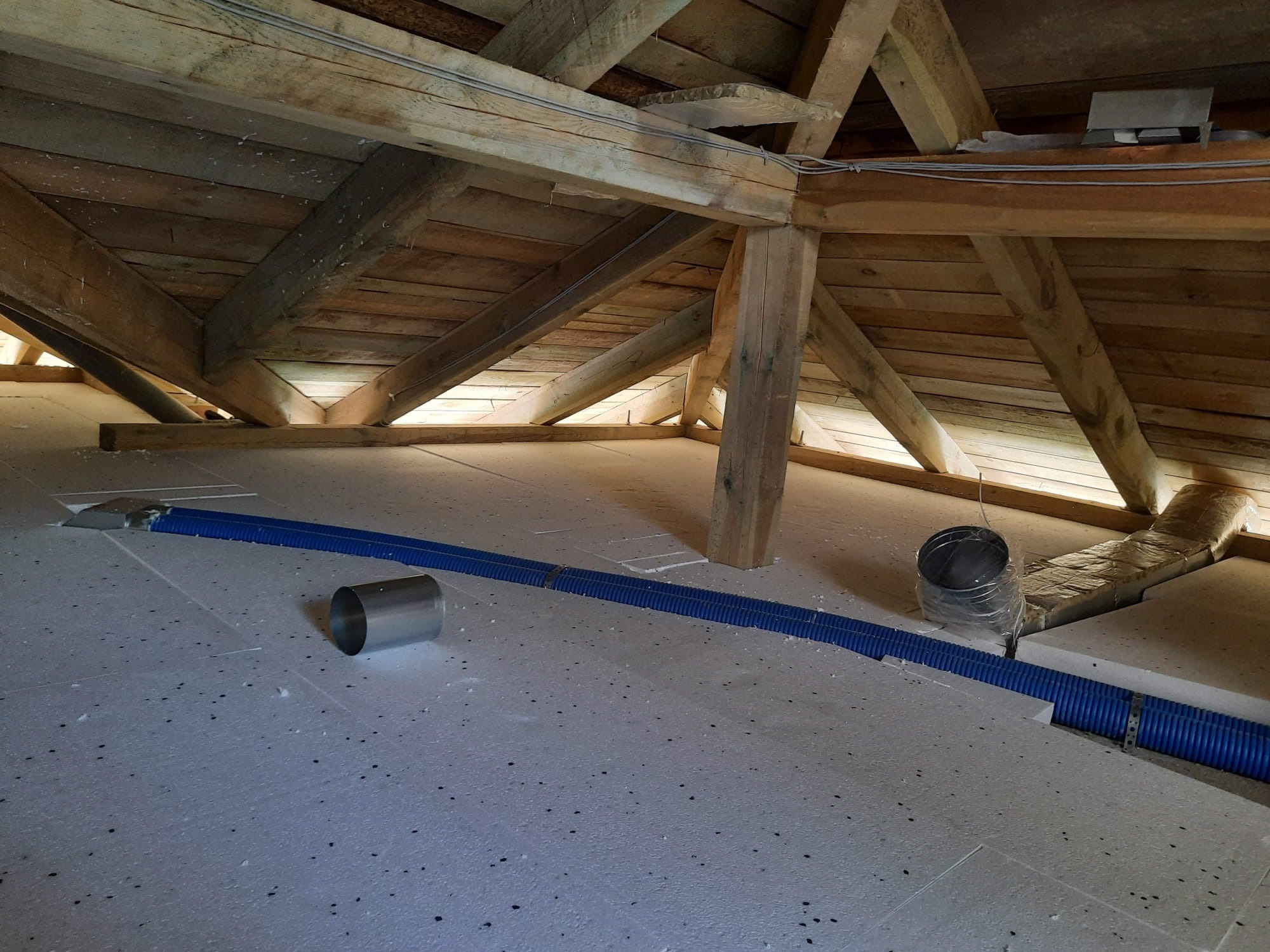 Poddasze z widoczną drewnianą konstrukcją dachu, pokryte białymi płytami izolacyjnymi z czarnymi kropkami i niebieskim przewodem wentylacyjnym oraz metalowymi elementami wentylacyjnymi.