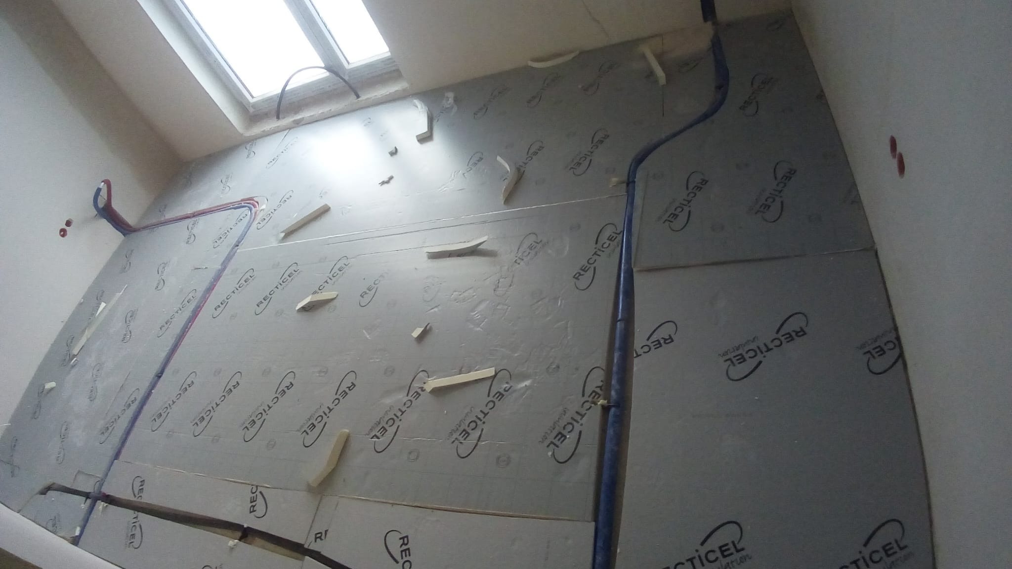 Wnętrze w trakcie prac budowlanych: ściana pokryta szarymi panelami izolacyjnymi z widocznym logo producenta 'RECTICEL', przymocowanymi drewnianymi klinami, z fragmentami instalacji wodnej...