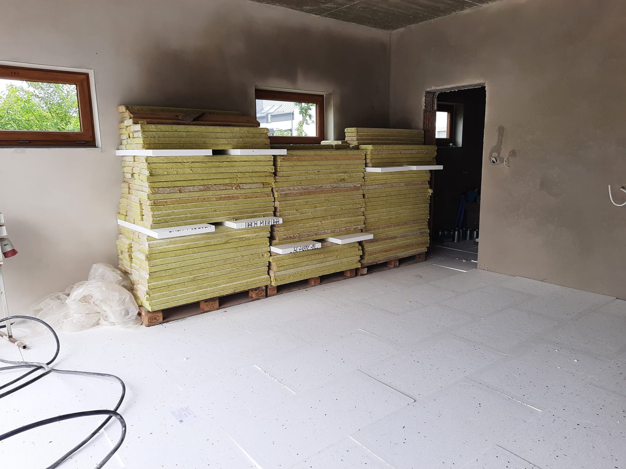 Stos płyt izolacyjnych z wełny mineralnej na paletach w surowym wnętrzu, widoczne okno i przygotowana podłoga z płyt styropianowych.