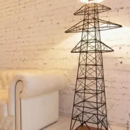 Lampa podłogowa w kształcie stylizowanej wieży energetycznej, z abażurem w ciepłym odcieniu, stojąca obok kremowej sofy na tle ściany z białej cegły.