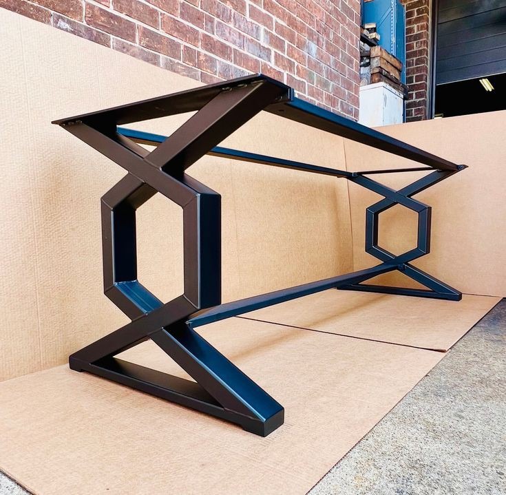 Metalowa podstawa stołu o geometrycznym wzorze, czarny mat, na tle ceglanej ściany i kartonowego podłoża.