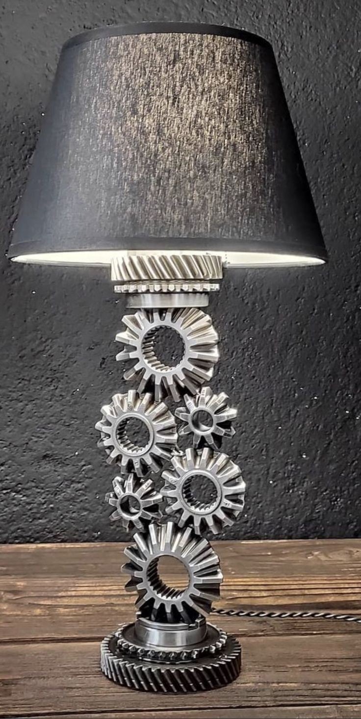 Lampa stołowa wykonana z metalowych kół zębatych w różnych rozmiarach, z czarnym abażurem, stojąca na drewnianym blacie na tle czarnej ściany o chropowatej fakturze.