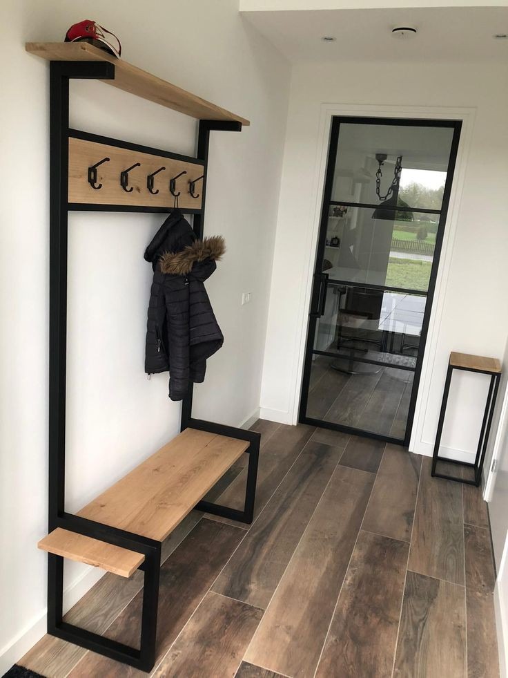 Nowoczesny przedpokój z drewnianą ławką, wieszakiem i półką na metalowej konstrukcji, kurtka zimowa wisi na haczyku, w tle przeszklone drzwi w czarnej ramie.