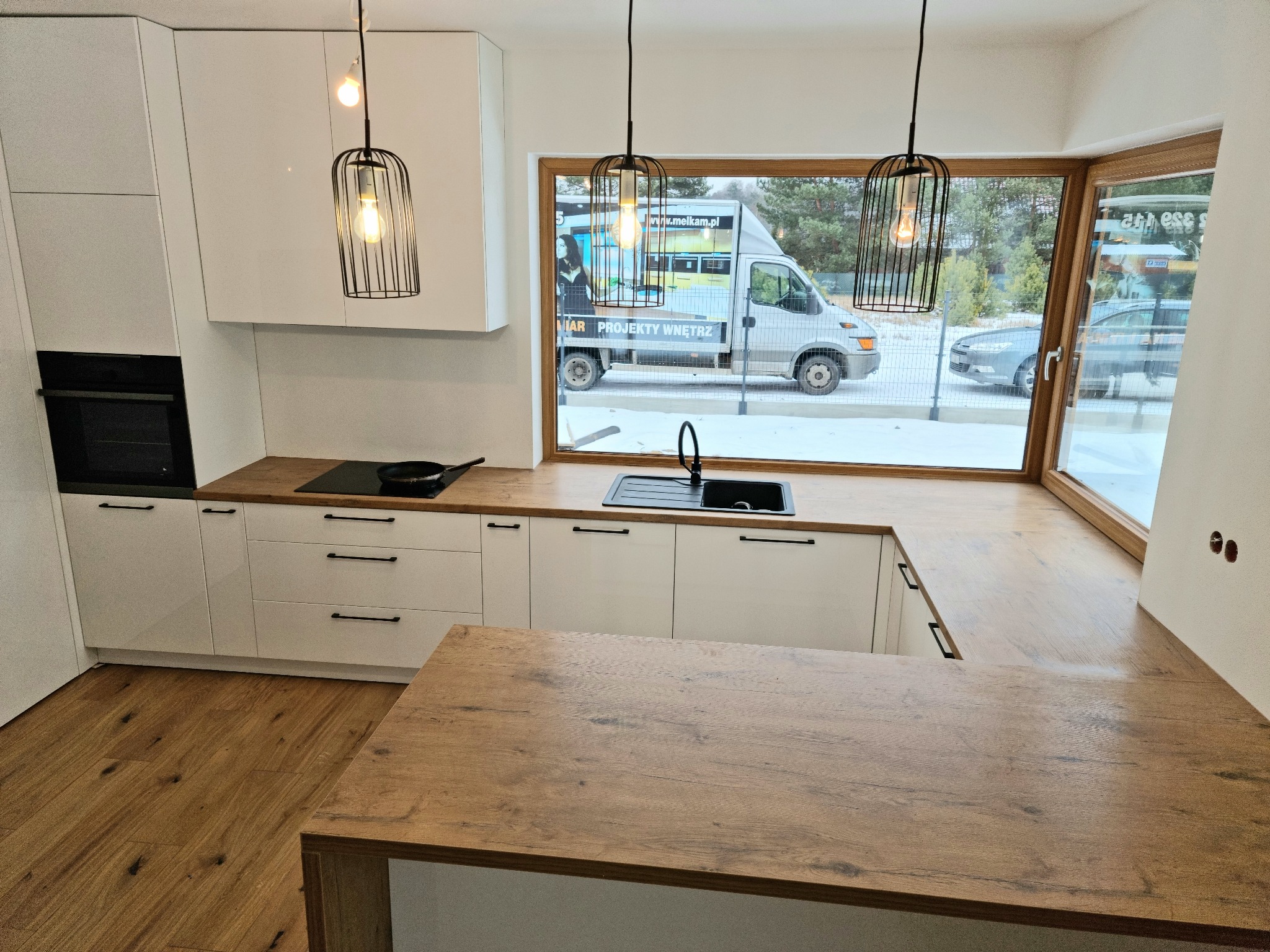 Nowoczesna kuchnia z białymi szafkami, drewnianym blatem i wyspą, oświetlona wiszącymi lampami w stylu industrialnym, z widokiem na firmowy samochód za oknem.