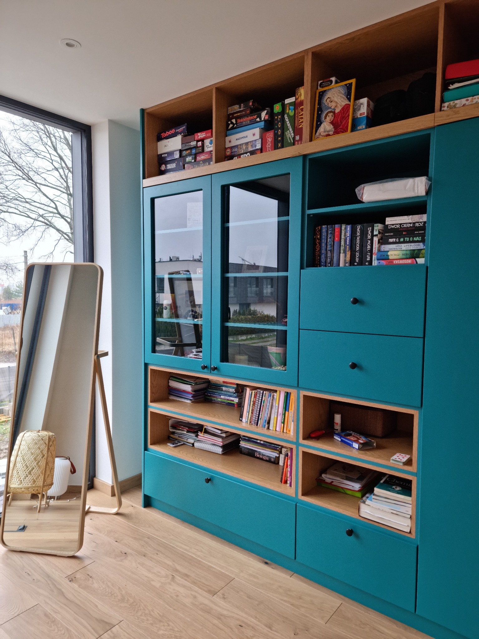 Turkusowa zabudowa meblowa z drewnianymi akcentami, zawierająca przeszklone witryny, otwarte półki z książkami i grami planszowymi oraz szuflady, uzupełniona o lustro stojące w drewnianej ramie...