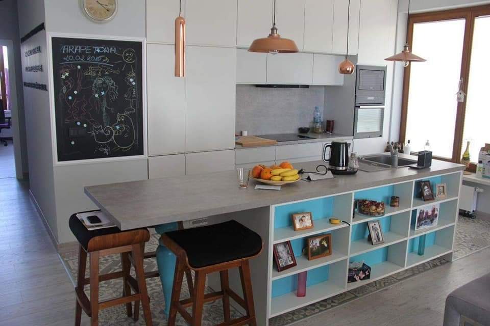 Nowoczesna, jasna kuchnia z wyspą i hokerami, ściana z tablicą kredową z rysunkami dziecięcymi, wiszące lampy w kolorze miedzi.