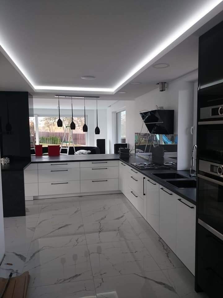 Nowoczesna kuchnia z białymi szafkami i czarnymi blatami, marmurowa podłoga, oświetlenie LED podsufitowe i wiszące lampy w tle, widok na ogród przez okno.